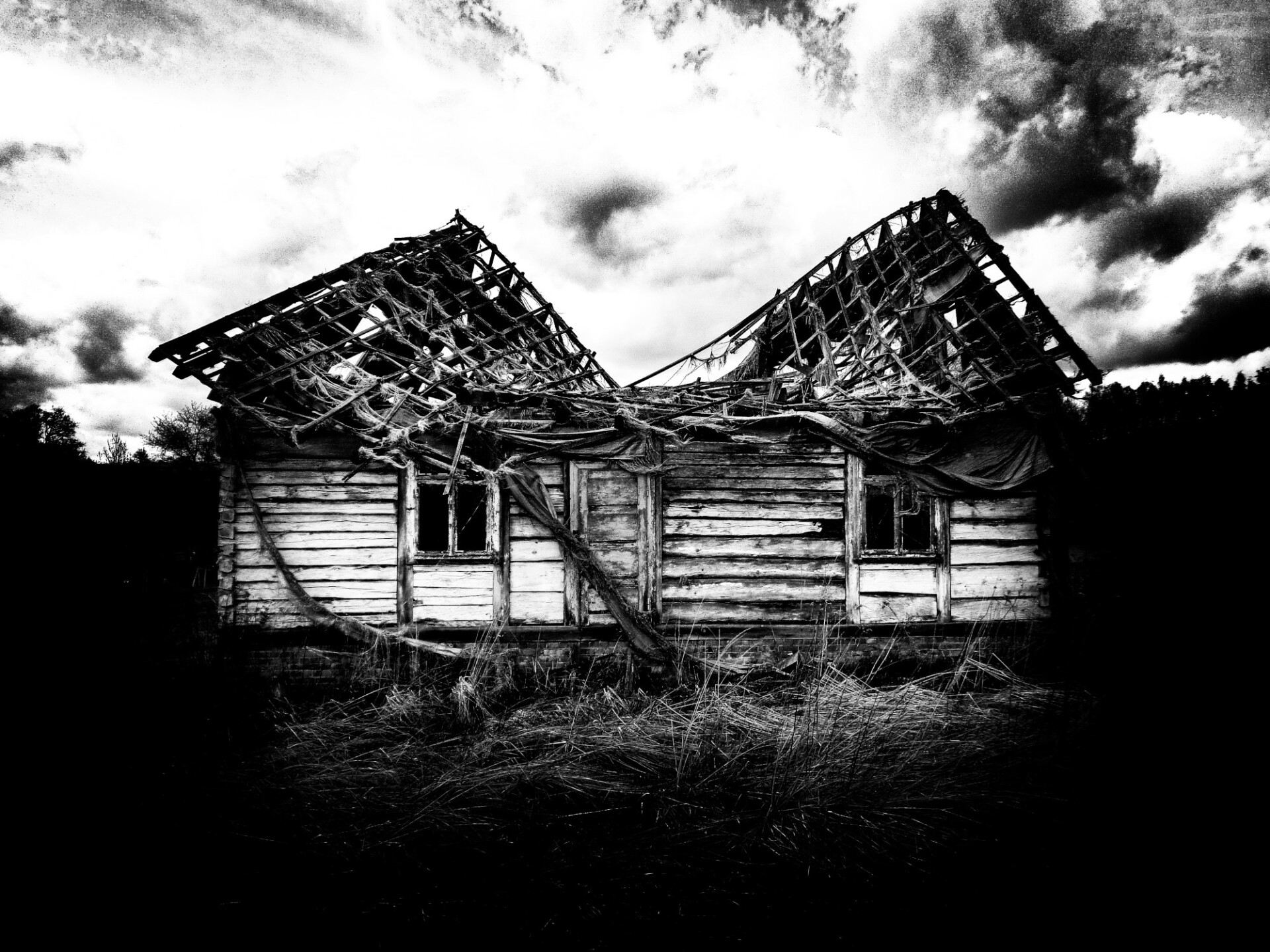 Czarno biała fotografia przedstawia drewniany stary dom z zawalającym się dachem na tle lasu i białych obłoków.