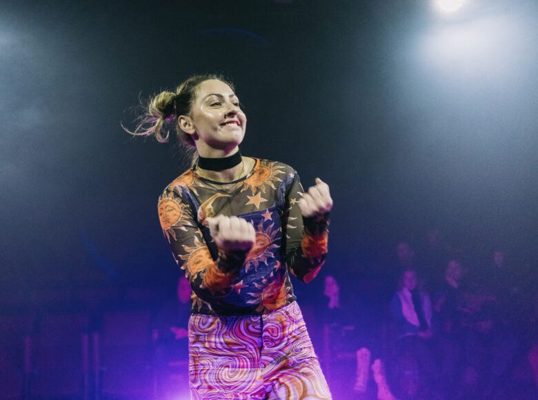 Zdjęcie przedstawia kobietę w kolorowym stroju, która stoi na scenie i uśmiecha się. Jeden róg sceny oświetla duży reflektor. Na fotelach widowni siedzi grupa ludzi.