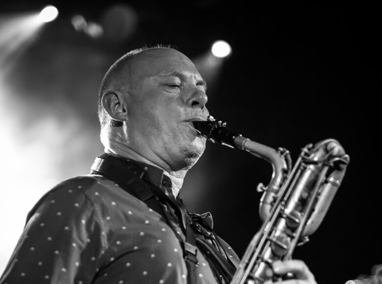 Na fotografii zbliżenie na mężczyznę grającego na saksofonie. Za nim ciemne tło z kilkoma punktami światła.