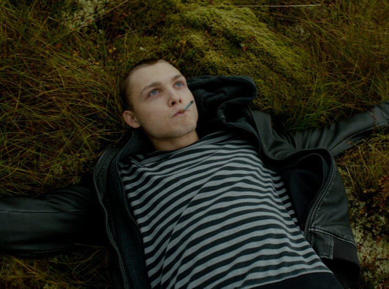 Młody chłopak z papierosem w ustach leży na trawie z rozłożonymi rękami w geście wolności. Ubrany jest w pasiasta koszulkę i czarną kurtkę.