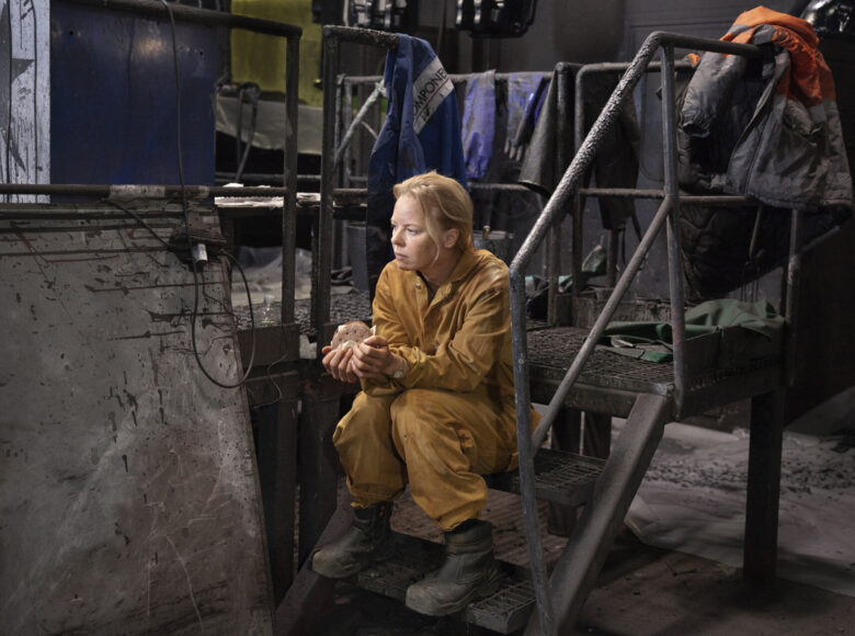 Kobieta w żółtym kombinezonie i zielonych kaloszach siedzi na schodach w fabryce. W rękach trzyma kanapkę. Jest zamyślona.