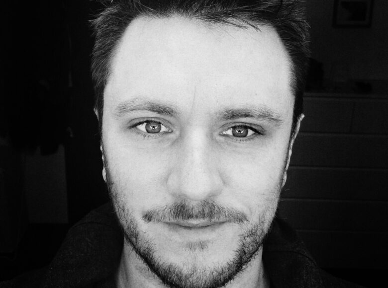 Czarno-biała fotografia. Na czarnym tle twarz młodego mężczyzny o czarnych włosach, które wtapiają się w czarne tło fotografii. Mężczyzna ma delikatny, rzadki, czarny zarost (wąsy i brodę), przez który prześwituje skóra. W szarych oczach patrzących z fotografii na widza, można dostrzec małe, błyszczące bliki światła.