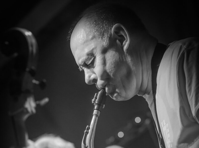 Na fotografii zbliżenie na mężczyznę grającego na saksofonie. Za nim ciemne tło z kilkoma punktami światła.