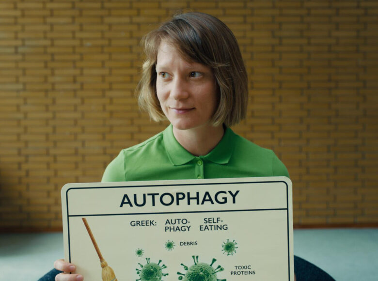 Kolorowa fotografia. Po środku kadru we wnętrzu siedzi młoda dziewczyna i trzyma przed sobą tabliczkę z napisem w języku angielskim: autophagy. Dziewczyna ma włosy do ramion i patrzy w prawą stronę z lekkim uśmiechem na ustach.