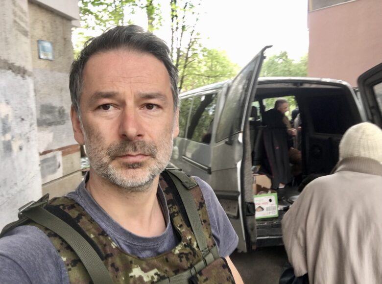 Kolorowa fotografia. Na pierwszym planie zbliżenie popiersia wizerunku męszczyzny z ciemnymi włosami i krótkim siwiejącym zarostem. Jest to autor wystawy Maks Skrzeczkowski, ubrany w szarą podkoszulkę z krótkim rękawem i wojskową kamizelkę kuloodporną z maskującym wzorem w panterkę. Na drugim planie widzimy niewielki szary samochód dostawczy z otwartymi podwójnymi tylnymi drzwiami oraz stojące przy nim 2 osoby ubrane w cywilne ubrania.