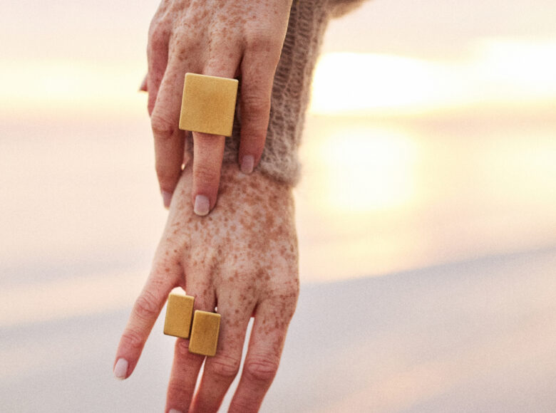 Kolorowe zdjęcie. Na zbliżeniu dwie kobiece dłonie. Kobieta nosi na ręce dużą, złotą bransoletę, a na palcach ma prostokątne pierścionki. Tło jasne, jednolite.