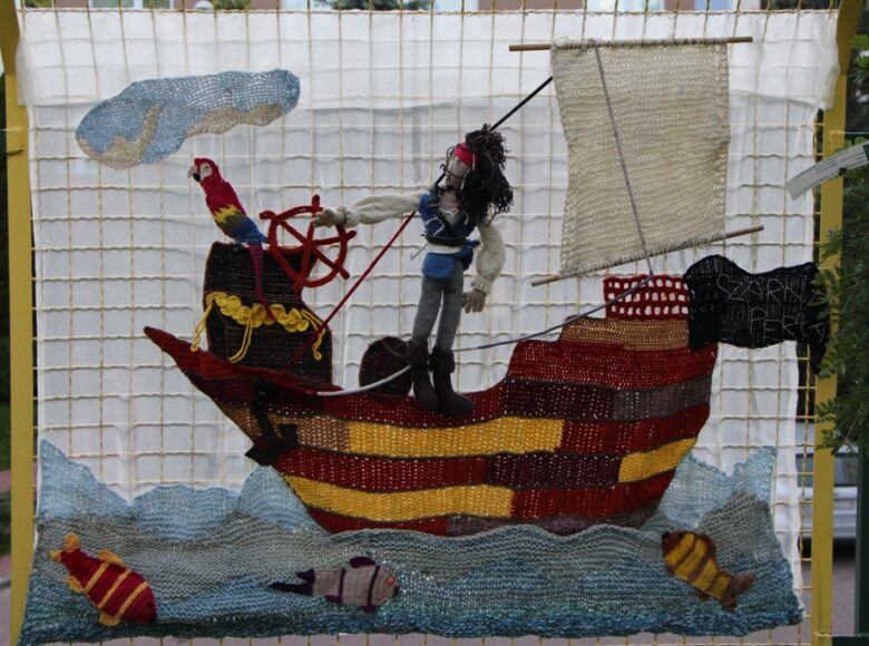 Zdjęcie przedstawia wyszyty włóczką obrazek, zawieszony na bramie. Obrazek przedstawia pirata płynącego statkiem.