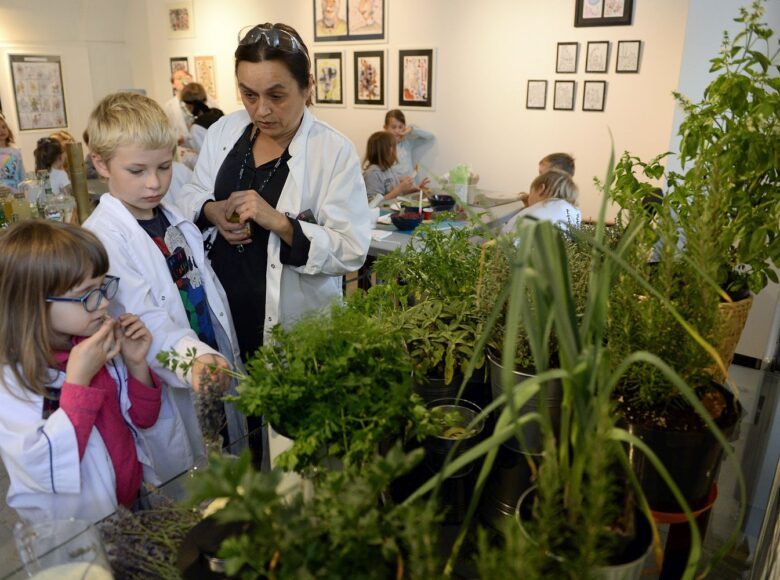 Kolorowe zdjęcie. Na pierwszym planie rośliny, zioła, dalej dwoje dzieci i jedna osoba dorosła w fartuchach laboratoryjnych.