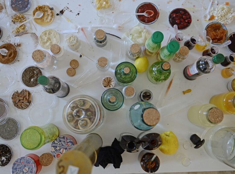 Zdjęcie przedstawia blat stołu, a na nim bardzo dużo naczynek laboratoryjnych z miksturami, próbkami jedzenia, wszystko w nieładzie.