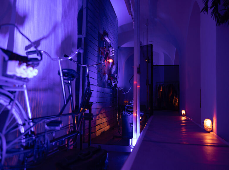 Zdjęcie przedstawia wnętrze, oświetlonego na fioletowo korytarza. Przy jednej ze ścian stoją stare rowery.