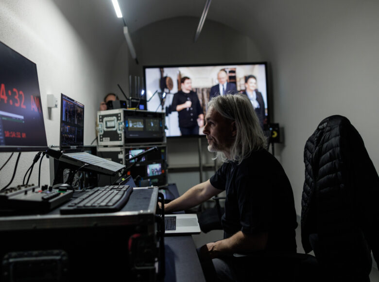 Zdjęcie przedstawia mężczyznę, który siedzi przed biurkiem. Na biurku stoją komputery i monitory. Na ścianie wisi telewizor.