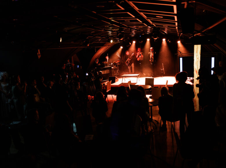 Zdjęcie przedstawia scenę, na której występują muzycy. Pomieszczenie jest oświetlony pomarańczowym światłem.