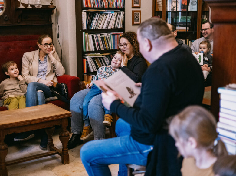 Wnętrze księgarni. Mężczyzna siedzi na krześle i czyta książkę. Na kanapach i fotelach siedzą rodzice z dziećmi i słuchają opowieści.