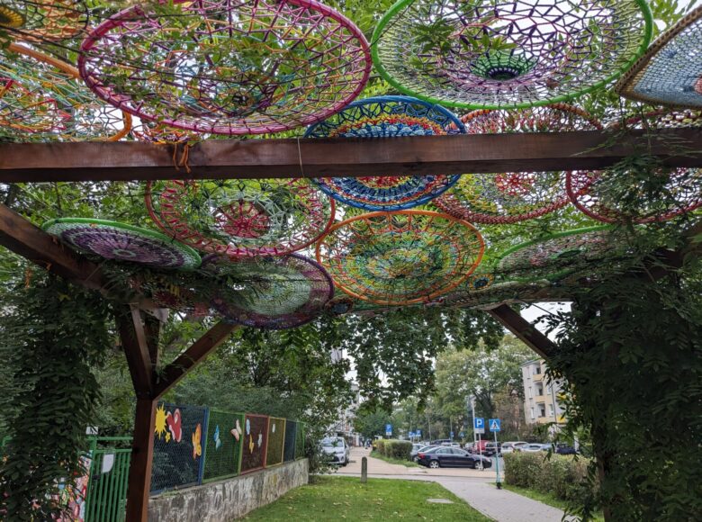 Zdjęcie przedstawia drewniana pergolę w miejskim parku. Na dachu pergoli zawieszone są kolorowe okręgi wykonane z włóczki.