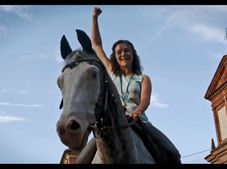 Kadr z filmu. Dziewczyna siedzi na koniu, jedną rękę trzyma wysoko w górze, uśmiecha się. W tle błękitne niebo i fasada kościoła.