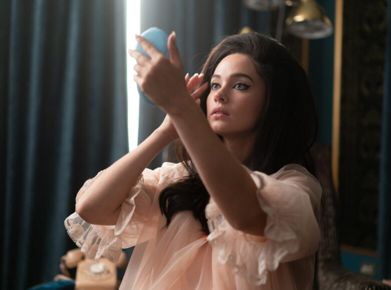 Kobieta ubrana w pudrowa podomkę, z fryzurą z lat 50. w pełnym makijażu przegląda się w pudernicy.