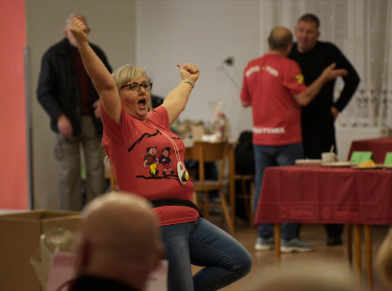 Kadr z filmu. Wnętrze. Kobieta w dżinsach i czerwonej koszulce szeroko rozkłada ręce wyciągnięte w górę. W rozmytym tle ludzie, stoły, kartony.