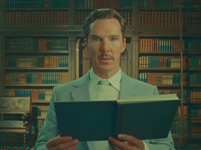 Kadr z filmu. Na tle ściany pełnej półek z książkami stoi mężczyzna w jasnej marynarce, który trzyma otwartą książkę i patrzy wprost na nas.