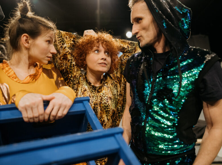 Kolorowe zdjęcie. Scena ze spektaklu. Obok siebie stoją trzy osoby w kostiumach. Aktorzy patrzą na siebie. Jedna z postaci w stroju żyrafy trzyma niebieski stołek.