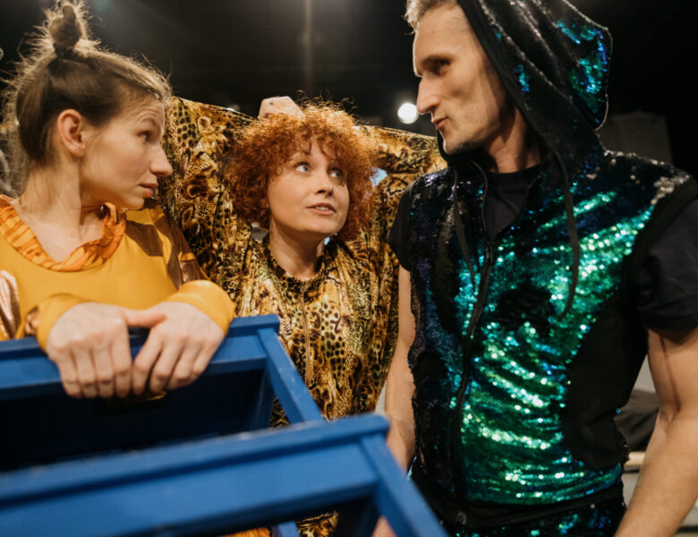 Kolorowe zdjęcie. Scena ze spektaklu. Obok siebie stoją trzy osoby w kostiumach. Aktorzy patrzą na siebie. Jedna z postaci w stroju żyrafy trzyma niebieski stołek.