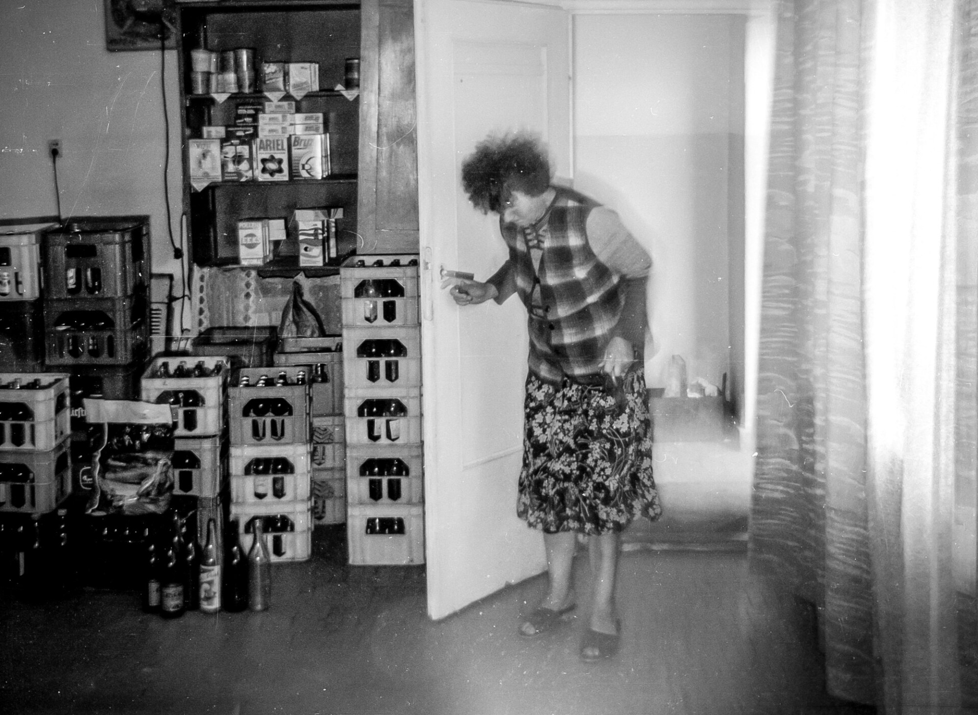 Fotografia przedstawia kobietę w średnim wieku, otwierającą drzwi i wchodzącą do wnętrza magazynu spożywczego sklepu. Świadczą o tym stojące na podłodze skrzynki z napojami i winem oraz półki z wystawionymi na nich artykułami spożywczymi.