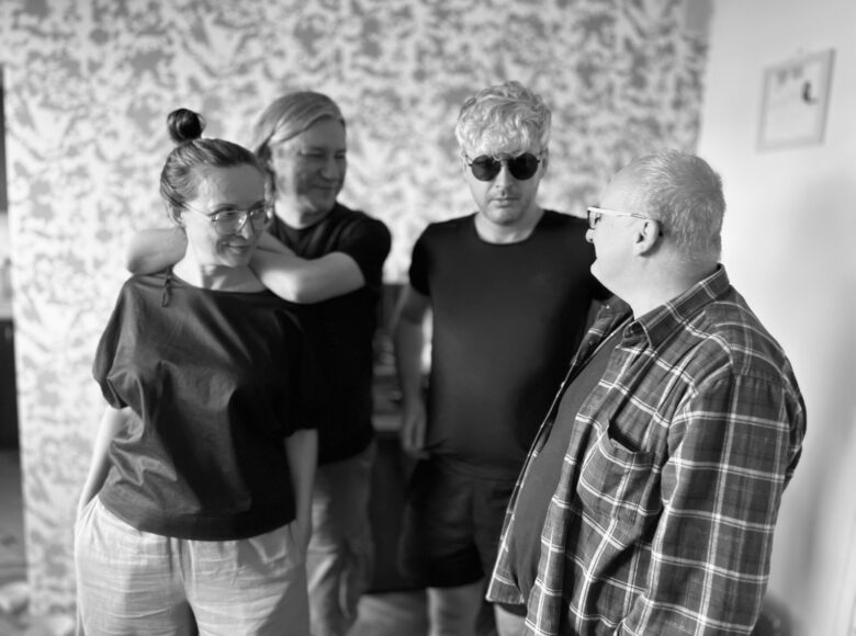 Czarno-białe zdjęcie. przedstawia trzech mężczyzn i kobietę. Grupa stoi na tle ściany pokrytej tapetą w kwiatki.