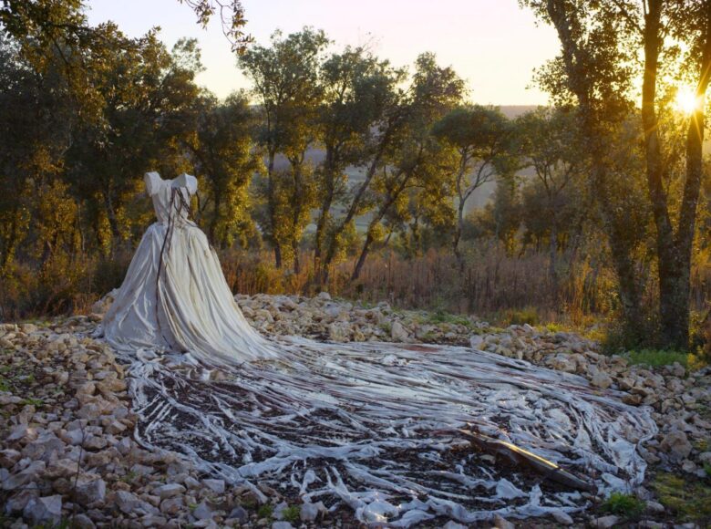 Kadr z filmu "Anselm". Polana otoczona drzewami. Na środku stoi manekin, na który nałożona jest biała, balowa suknia z trenem. Tren jest ubrudzony i lekko przysypany ziemią.
