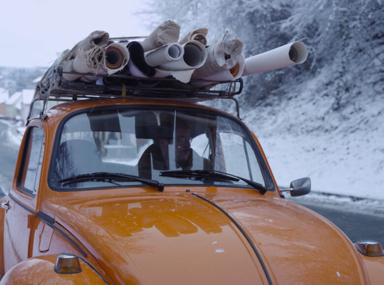 kadr z filmu "Anselm" Na zbliżeniu mały, pomarańczowy samochód. Na dachu samochodu zamocowano kilka zwiniętych rulonów tkanin. Samochód jedzie drogą w górach.