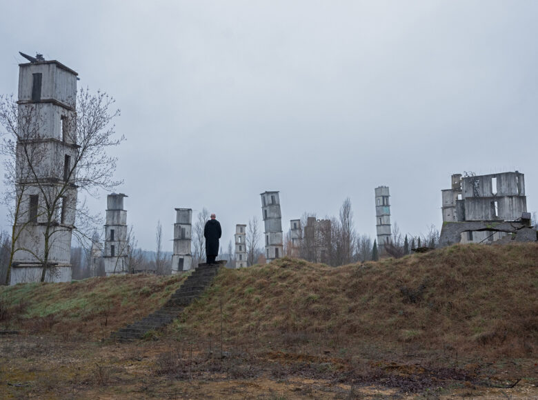 Zdjęcie przedstawia widok na wzgórze na którym rozstawione są monumentalne rzeźby przypominające muzy, blokowiska. Wśród rzeźb widoczna jest postać mężczyzny ubranego na czarno. Całość utrzymana w ponurej kolorystyce.