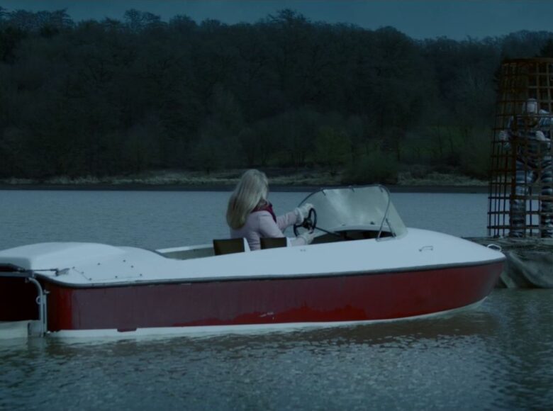 Kadr z filmu. Kobieta w motorowej łódce podpływa do platformy, na której znajduje się metalowa klata z uwiezionym w środku człowiekiem.