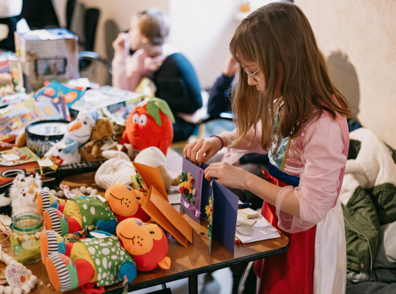 Zdjęcie przedstawia dziewczynę ubrana w sukienkę księżniczki. Dziewczyna stoi przy stole, na którym leżą kolorowe zabawki. Dziewczynka trzyma w rękach kolorowe kartki.