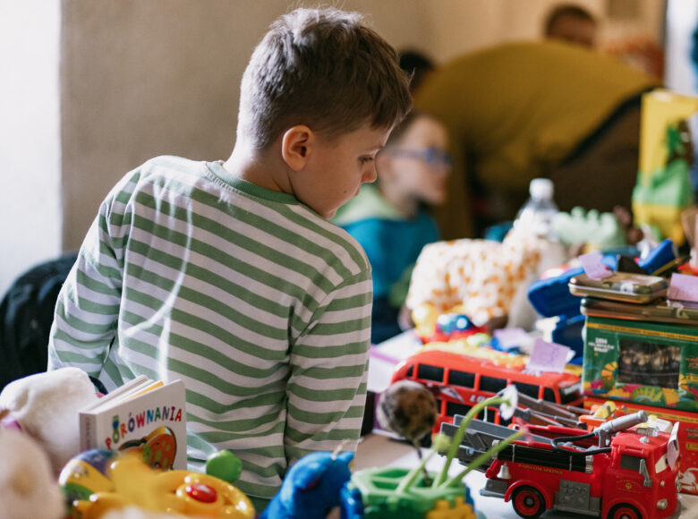 Zdjęcie przedstawia chłopca ubranego w koszulkę w biało-zielone paski. Chłopiec stoi obok stoiska pełnego zabawek, misiów i samochodzików.