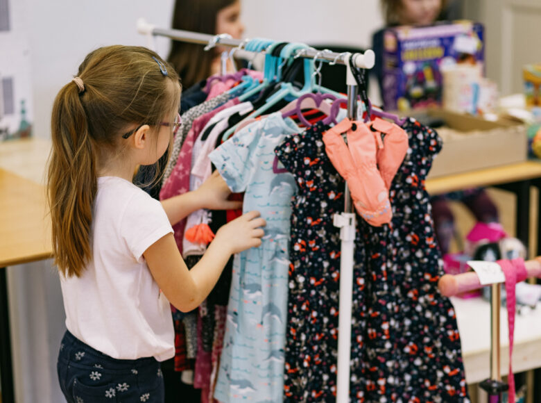 Zdjęcie przedstawia dziewczynkę, która przegląda wieszaki z kolorowymi sukienkami.