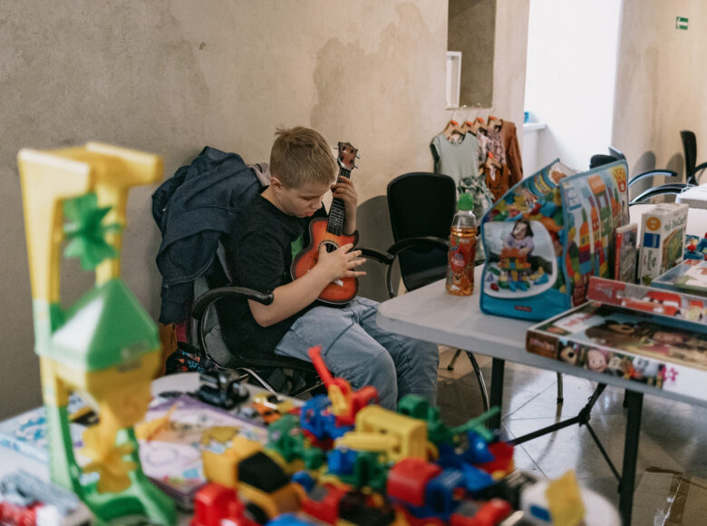 Zdjęcie przedstawia chłopca, który siedzi na krześle i gra na ukulele. Przed nim znajdują się stoły zastawione kolorowymi zabawkami, książeczkami, pudełkami kloców i puzzli.
