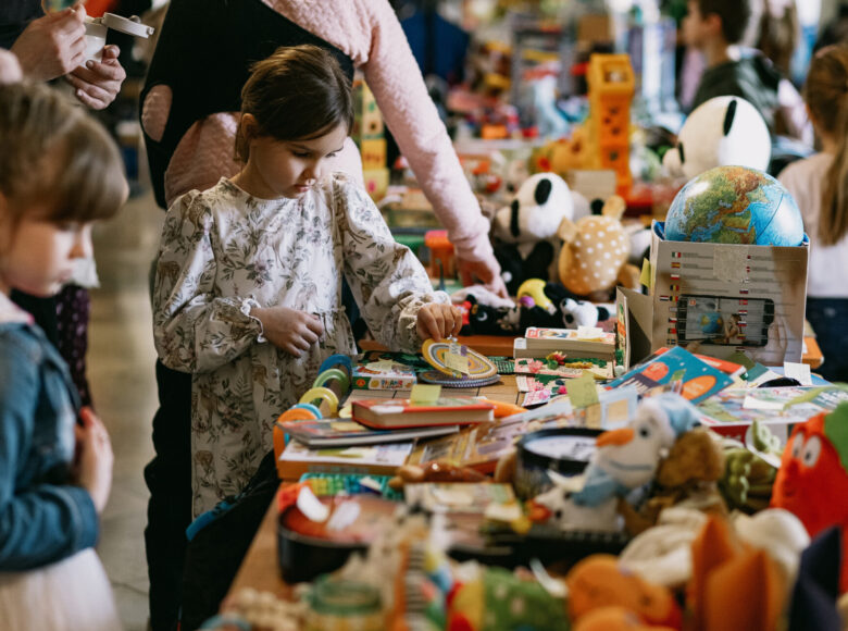 Zdjęcie przedstawia dwie dziewczynki, które stoją obok długiego stołu, na którym leżą kolorowe zabawki i książeczki.