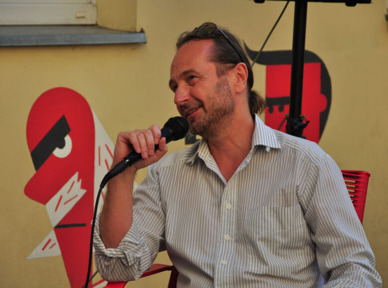 Zdjęcie przedstawia mężczyznę, który siedzi na krześle, w ręce trzyma mikrofon i mówi do niego.