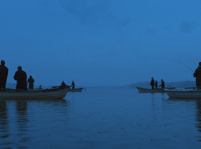 Kadr z filmu. Scena wieczorna. Na jeziorze znajduje się kilka łódek, a w nich rybacy, którzy łowią ryby. W oddali znajdują się góry i zabudowania .