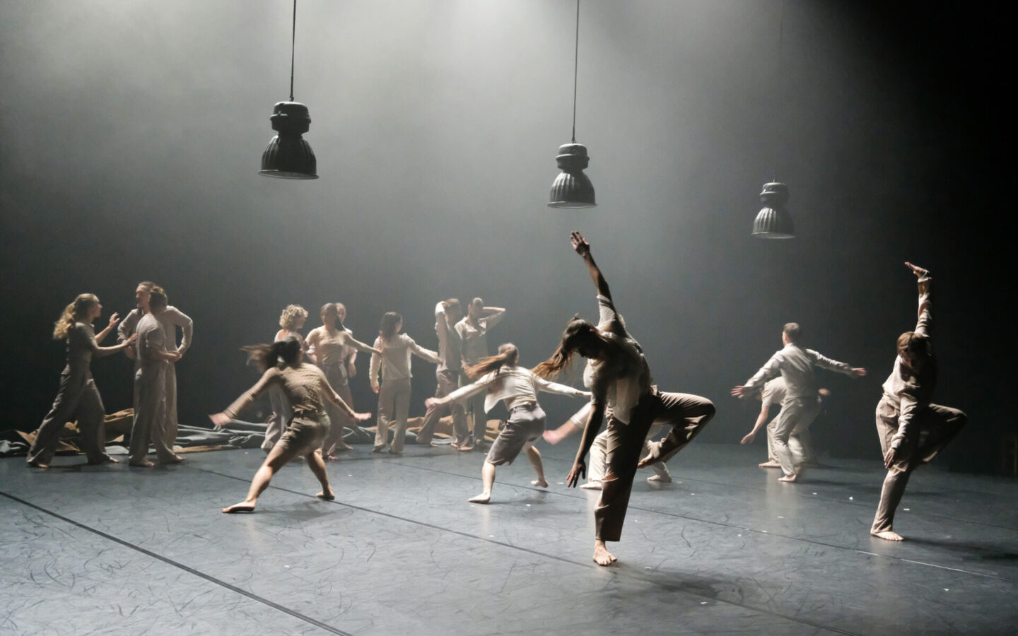 Kolorowa fotografia. Na scenie tańczy kilkanaście osób. Wszyscy ubrani są w luźne ubrania w odcieniach beżu. Białe światło oświetla tancerzy, widać unoszący się w powietrzu dym.