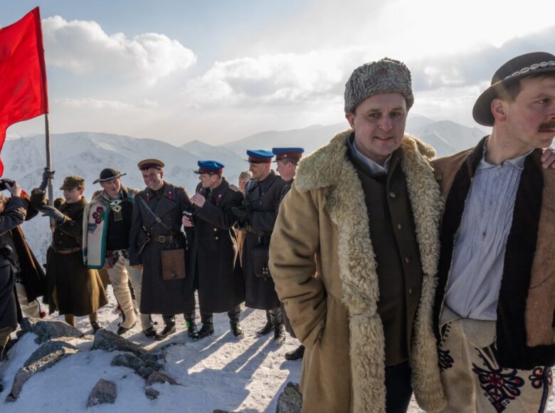 Na szczycie góry zgromadzeni są górale, którzy zadowoleni są z wspinaczki i osiągniętego celu. W tle powiewa czerwona flaga.