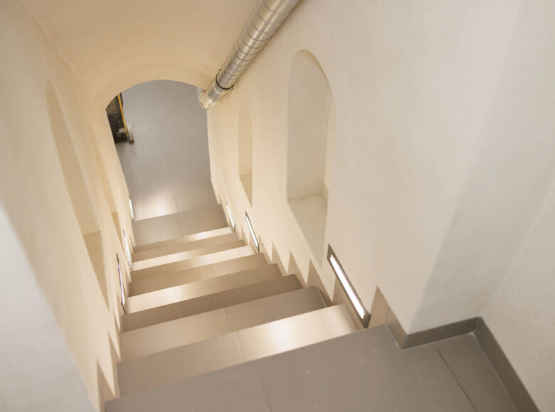 Zdjęcie przedstawia widok z góry klatki schodowej. Ściany są białe, a podłoga pokryta szarymi płytkami.