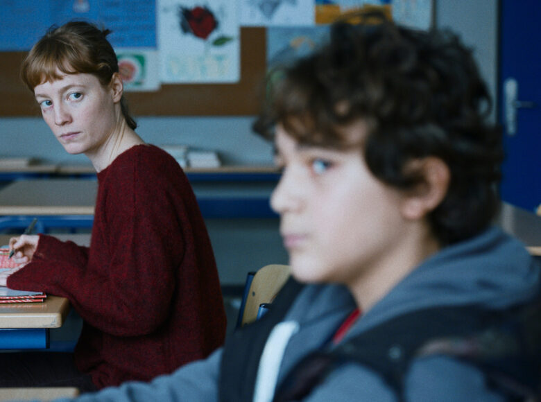 Zdjęcie przedstawia nauczycielkę i ucznia w szkole. Oboje siedzą w osobnych ławkach. Nauczycielka bacznie obserwuje ucznia. Uczeń jest zapatrzony i zamyślony, nie zwraca uwagi na otoczenie.