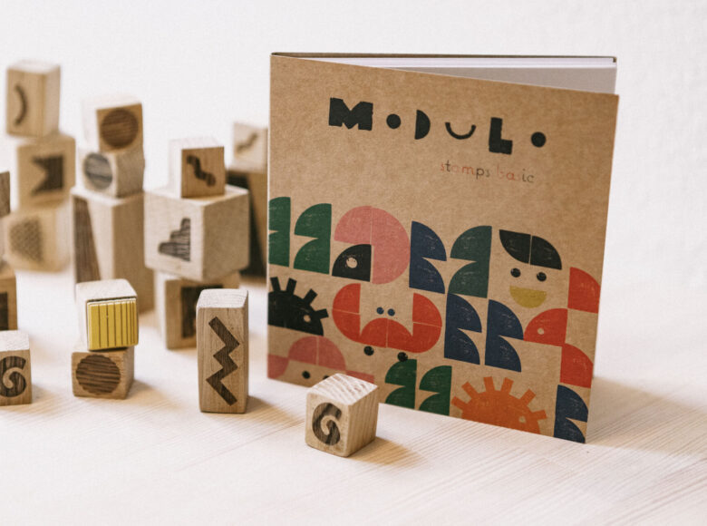 Zdjęcie przedstawia drewniane stemple z geometrycznymi wzorami. Obok znajduje się książka z wykonaną za ich pomocą kolorową grafiką.