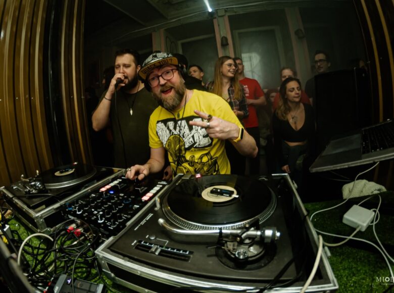 Zdjęcie z imprezy tanecznej. Na pierwszym planie gramofony i system nagłośnienia, przy których stoi DJ ubrany w żółtą koszulkę i czapeczkę z daszkiem. Uśmiecha się szeroko i unosi lewą rękę z palcami w geście Victoria. Obok niego stoi mężczyzna ubrany na czarno, śpiewa do mikrofonu. Za nimi tłum bawiących się, tańczących ludzi.