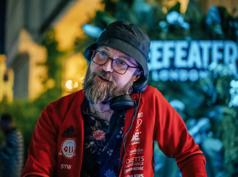 Zdjęcie mężczyzny. Ubrany jest w czarną koszulę w kwiaty, na wierzchu czerwoną bluzę z białymi napisami w języku angielskim. Mężczyzna ma brodę i nosi okulary. Na głowie ma ciemnozieloną czapeczkę typu wędkarskiego. Pomiędzy lewym uchem a lewym ramieniem przytrzymuje duże słuchawki nauszne. W tle niewyraźna ściana budynku i zielone rośliny.