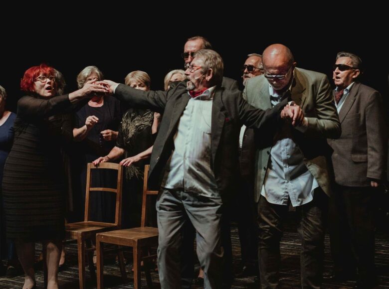 Scena ze spektaklu. Na scenie znajduje się grupa seniorów. Wszyscy patrzą na mężczyznę, który stoi na środku.