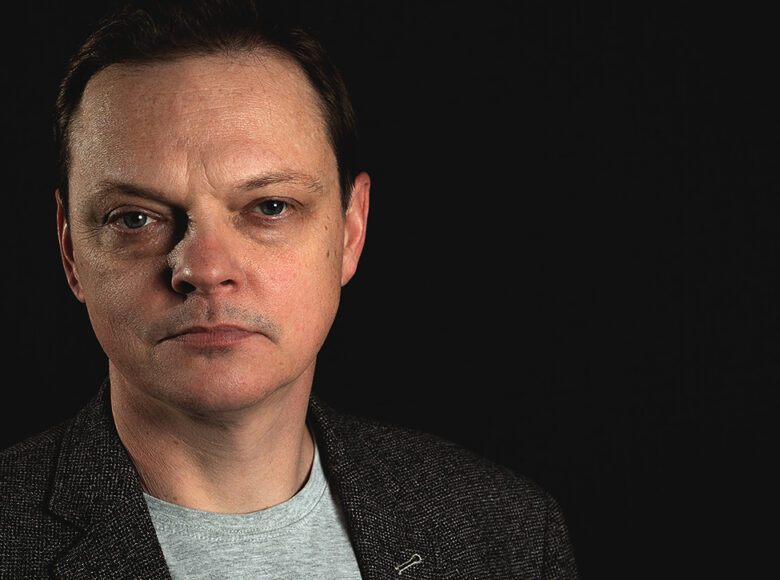 Zdjęcie. Portret mężczyzny. Mężczyzna patrzy przed siebie, ma poważny wyraz twarzy. Tło ciemne, jednolite.