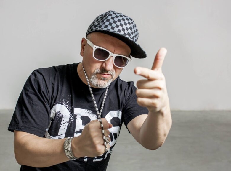 Zdjęcie mężczyzny. Ubrany jest w czarny t-shirt z białym napisem litery OBS. Wyciąga rękę w geście pistoletu. Drugą ręką ściska metalowy naszyjniki. Mężczyzna ma krótką siwą brodę. Ma okulary przeciwsłoneczne o białych oprawkach. Na głowie ma czapeczkę z daszkiem. Apteczka jest w biało-czarną kratę. Tło jest jasne – betonowa podłoga i jasna ściana.