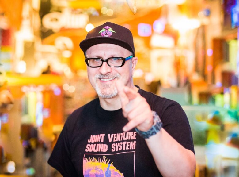 Zdjęcie mężczyzny. Ubrany jest w czarny t-shirt z różowym napisem w języku angielskim „Joint Venture Sound System”. Jedną ręką wyciąga do przodu w geście pozdrowienia, ma na niej masywny sportowy zegarek. Mężczyzna ma krótką siwą brodę i okulary w czarnych oprawkach. Na głowie ma czarną czapeczkę z daszkiem. Tło rozmazane, kolorowe, żółte i fioletowe światła.