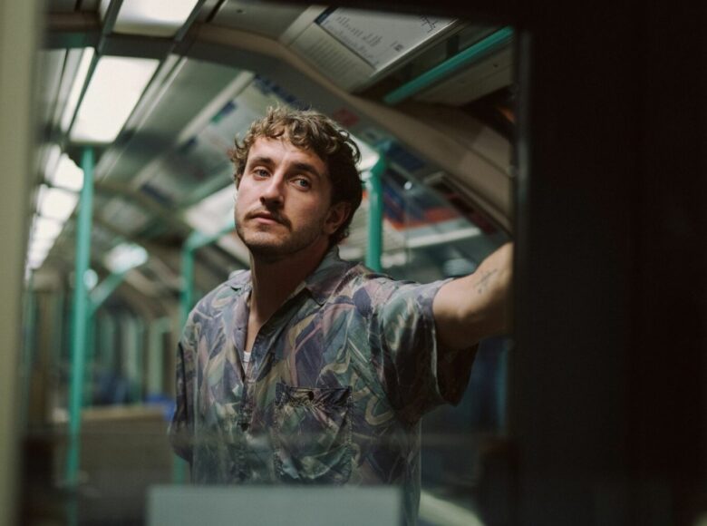 Kadr z filmu. Młody mężczyzna stoi w wagonie pociągu. Ubrany w kolorową koszulę z krótkimi rękawami, patrzy przed siebie.