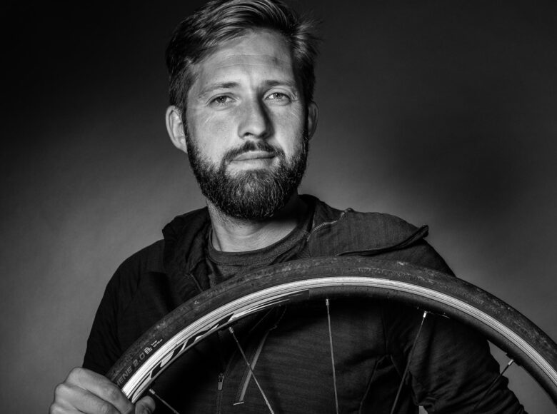 Czarno-białe zdjęcie. Portret mężczyzny. Jest ubrany w bluzę, uśmiecha się. W rękach trzyma koło od roweru.